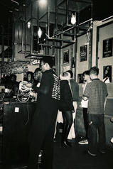 A greyscale photograph of a crowd of people in a small bar surrounded by pictures on the wall.
