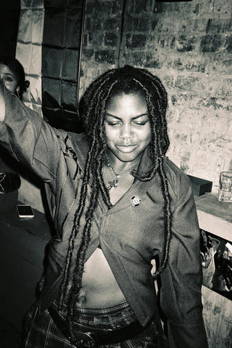 A greyscale photograph of a Black woman with long locs facing forward singing. She is in front of a brick wall.