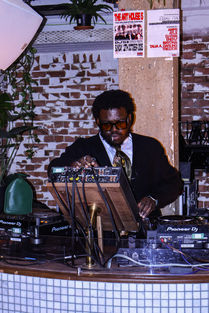 A black man who is wearing dark sunglasses, has a short afro, a lollipop in his mouth and is wearing a black cardigan with a sating gold and green tie and a white shirt. He is djing.