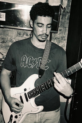 A greyscale photograph of a mixed race man wearing an 'AC DC' t-shirt and he is playing a white guitar. There is a brick wall in the background.