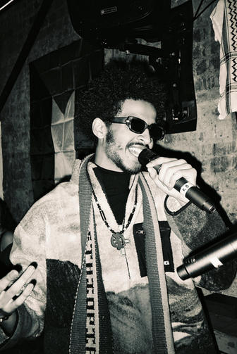 A greyscale photograph of a man with Afro hair singing with a microphone in his hand and he is wearing dark sunglasses and a light coloured fluffy coat. In the background is a brick wall.