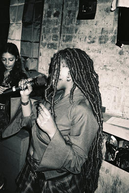 A greyscale photograph of a Black woman with long locs singing with a microphone in her hand. Her face is slightly concealed by her hair and there is a brick wall in the background behind her.