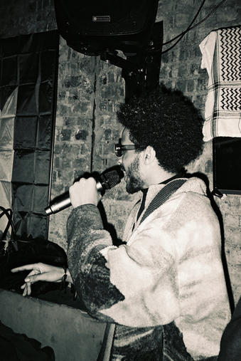 A greyscale photograph of a man with Afro hair and dark sunglasses singing with a microphone in his hand and he is stood sideways. There is a brick wall in the background.