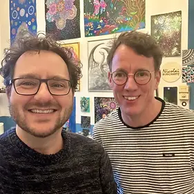Nicolai Burchartz und Tom schauen in die Kamera und lächeln. Im Hintergrund hängen viele gemalte Bilder an der Wand.