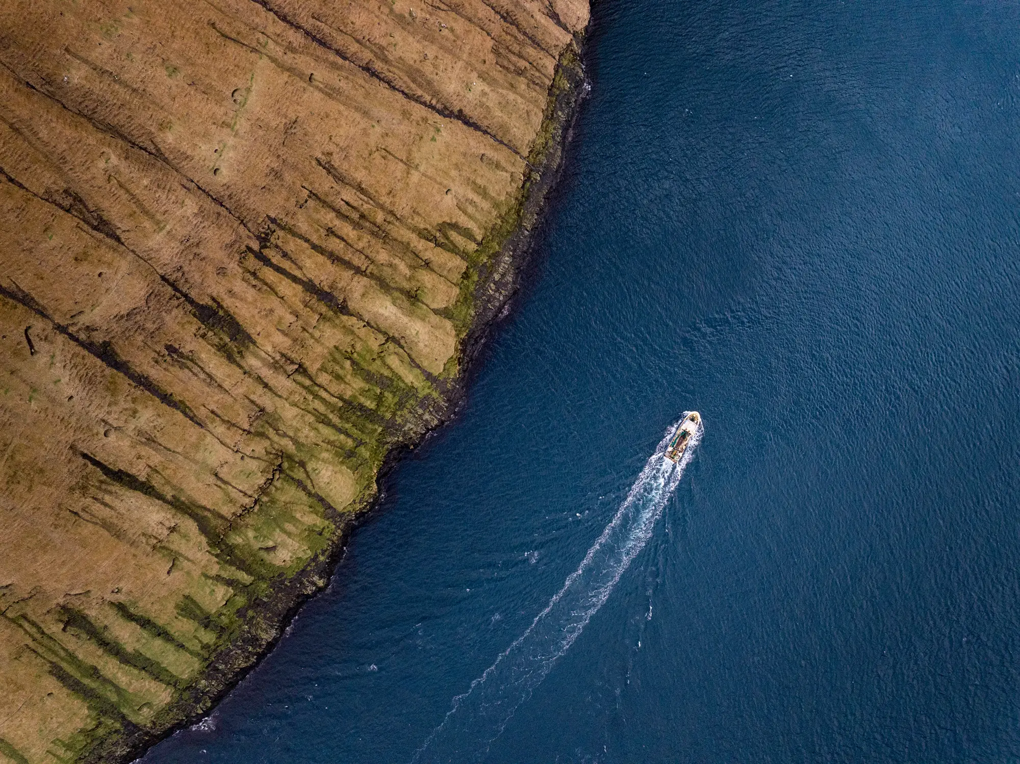 Ein Boot zieht seine Spur durch das tiefblaue Wasser entlang der steilen, grasbedeckten Klippen der Färöer-Inseln.