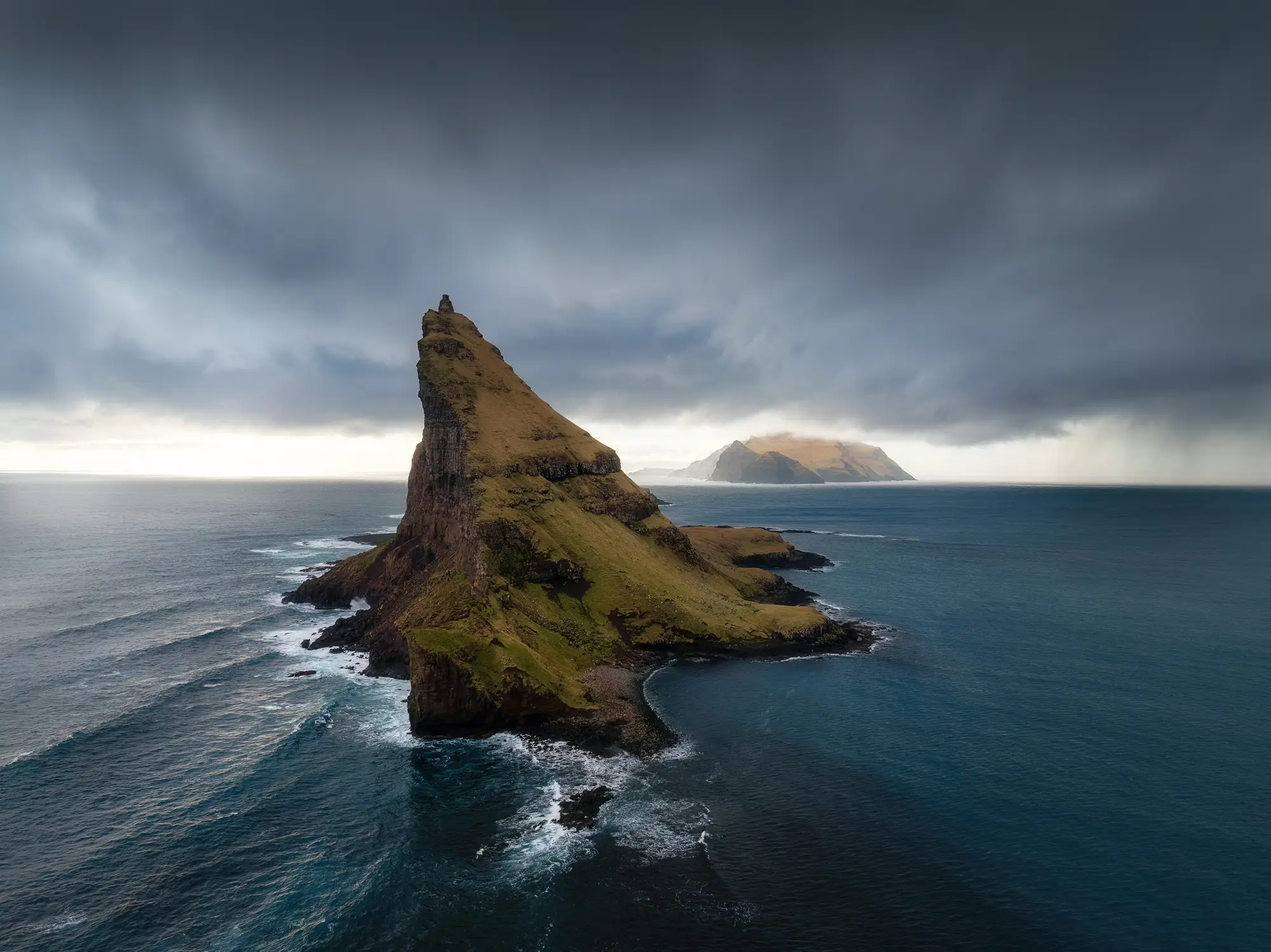 Eine markante Felseninsel ragt aus dem Atlantik, umgeben von dramatischen Wolken und wogenden Wellen der Färöer-Inseln.