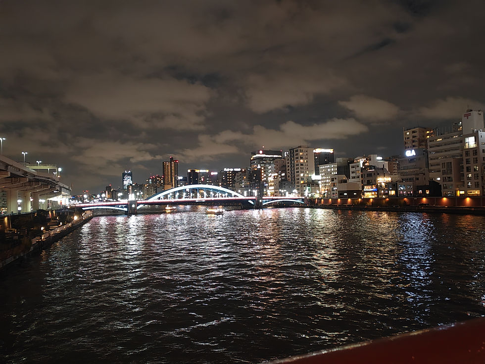 Night view across the dark waters of the Sumida River in Tokyo looking toward the illuminated white arches of Kachidoki Bridge.