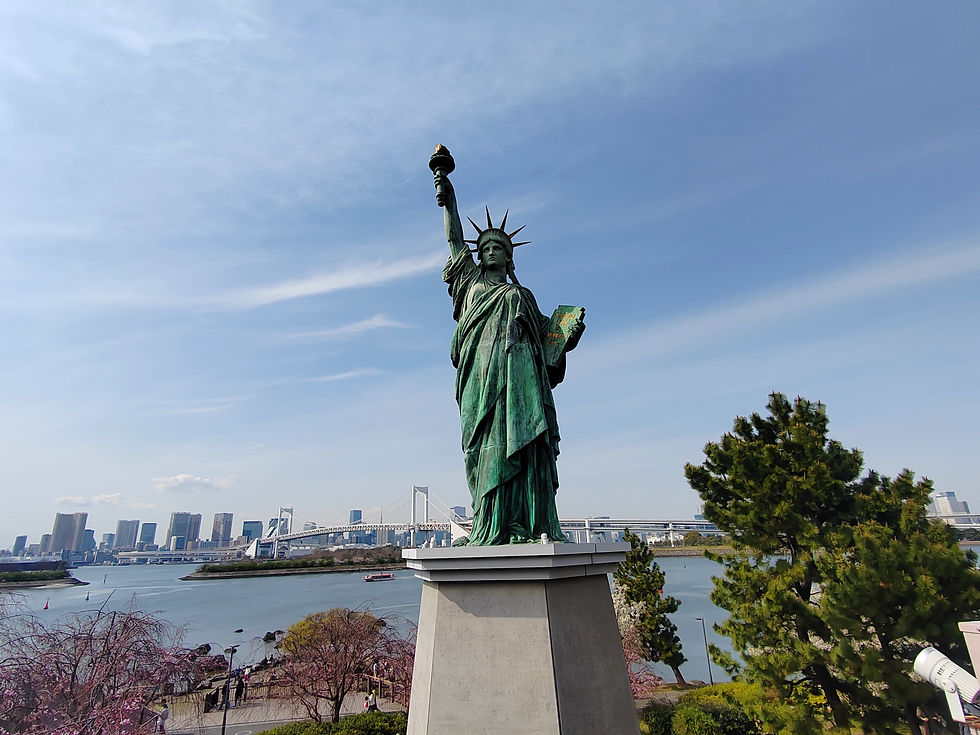 Bronze replica of the Statue of Liberty in Odaiba Marine Park with Tokyo Bay and cherry blossoms in the background.