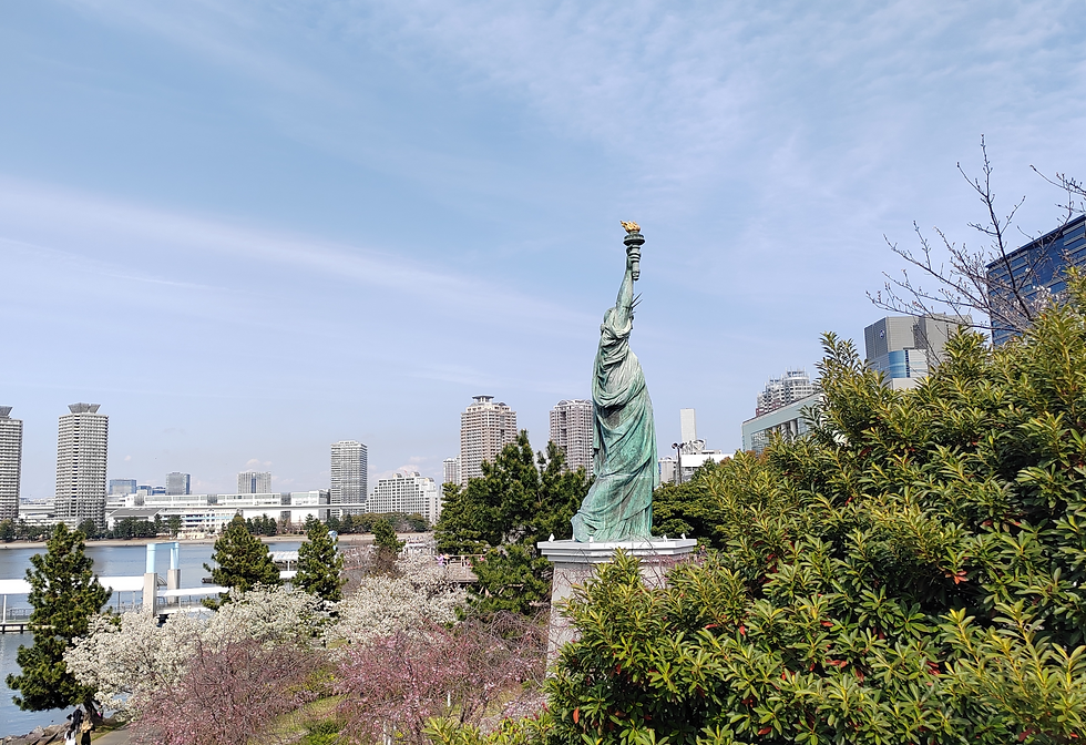 Bronze replica of the Statue of Liberty in Odaiba Marine Park with Tokyo Bay and cherry blossoms in the background.
