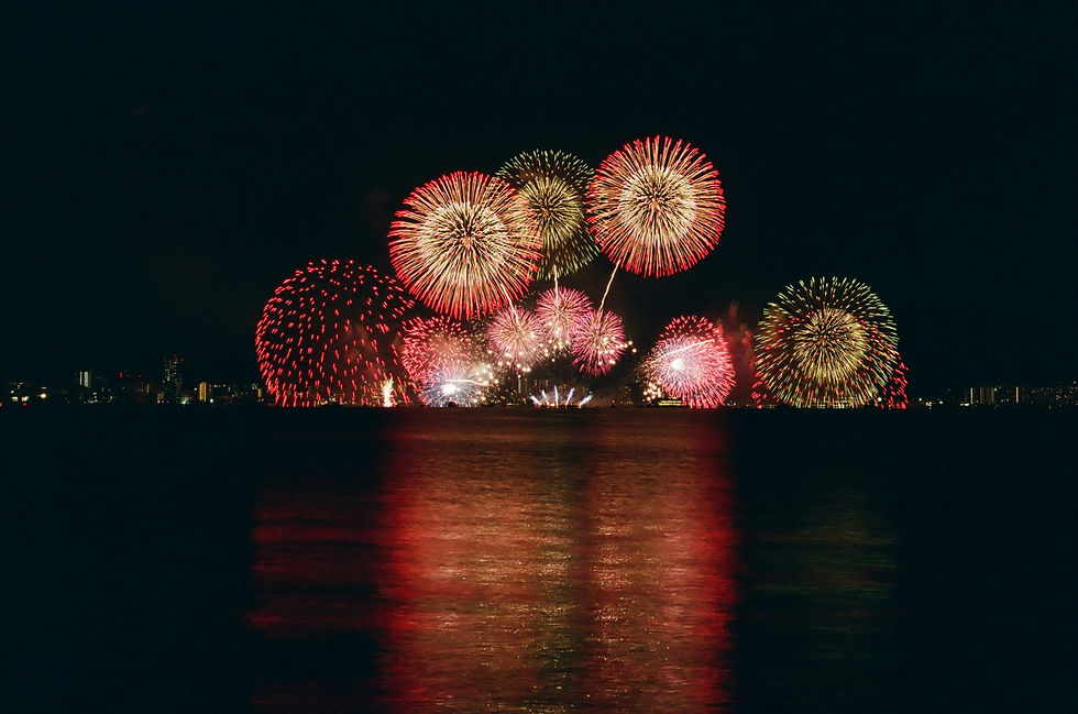 Colorful fireworks burst over a dark waterfront city skyline, with red, gold, and pink reflections on the water at night Japan fireworks and summer festivals while traveling japan for tourism.
