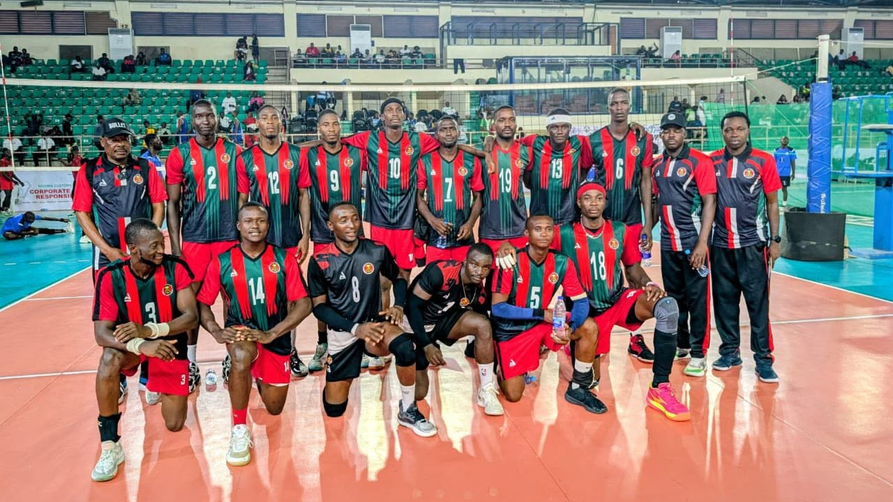 Chief of Army Staff (COAS) Volleyball team poses on an indoor court in red, green, and black jerseys, with coaches and spectators behind them.