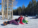 Two kids clipped into snowboards and wearing snow suits take a break to sit on the snowy mountain, the ski lift runs overhead behind them.