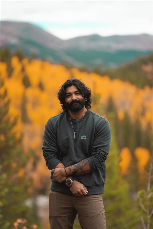 Kaartik has brown hair and a brown beard. He is in focus; behind him, a sea of orange trees is blurred.