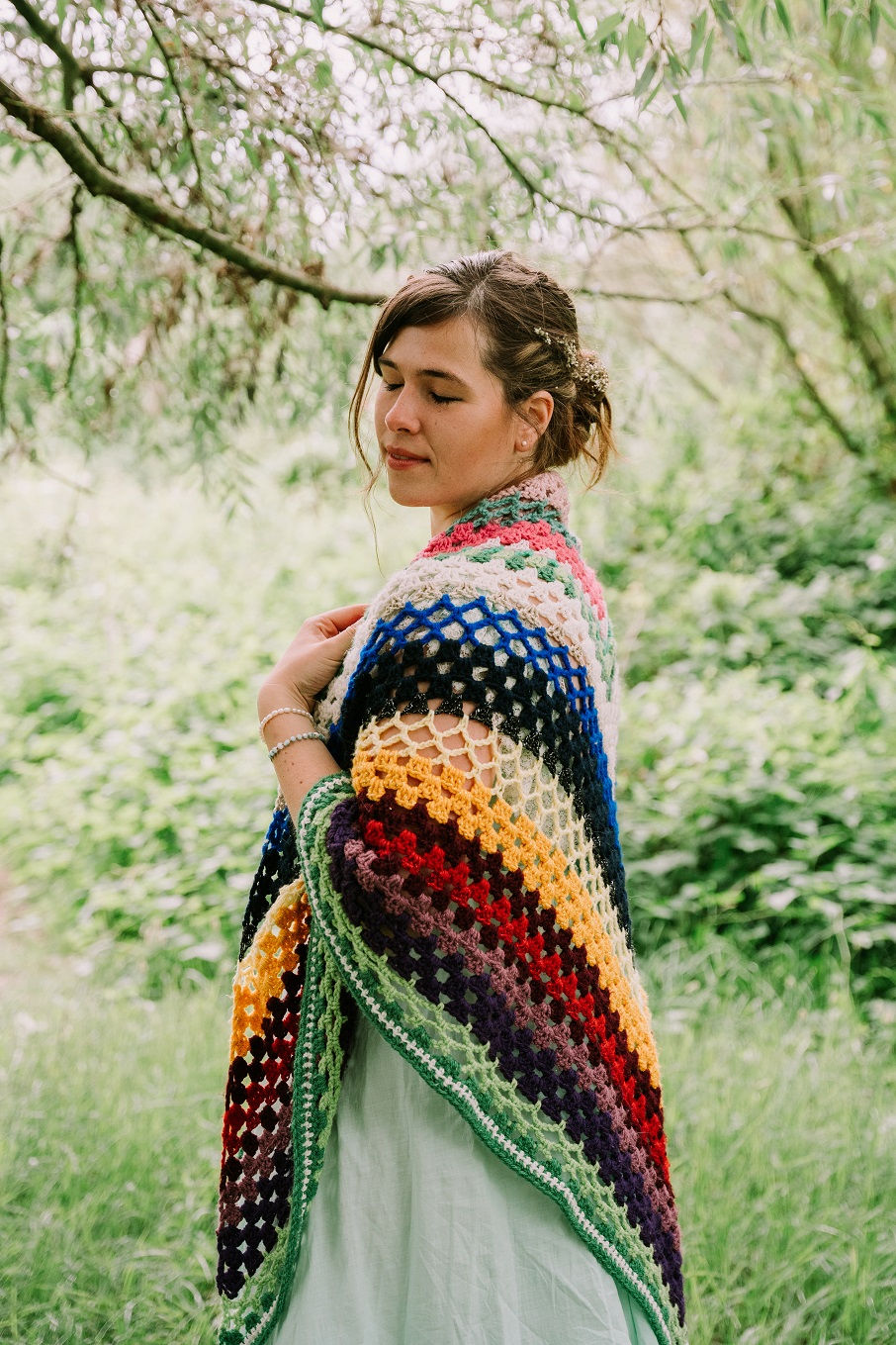 Vrouw in tuin met dichte ogen, draagt kleurrijke gehaakte omslagdoek. Rustige sfeer, groene achtergrond met loofbomen.