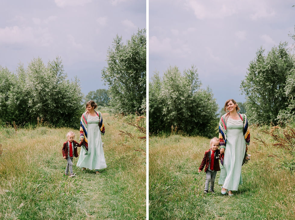 Vrouw in groene jurk en kleurrijk vest wandelt met kind in rood jasje door groen veld. Blauwe lucht, bomen op achtergrond. Beiden lachen.