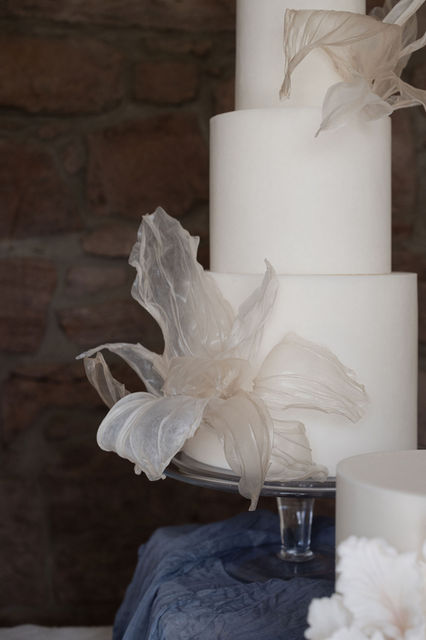 Three-tier sculptural wedding cake with minimal rice paper detailing, set against a raw Highland stone wall in a luxury Scottish venue.