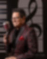 Marc Toussant, Jazz Singer, smiles broadly in a jacquard diiner jacket with a red pocket square. He holds a retro style microphone in front of Songbird Live's Piano Key stage backdrop.