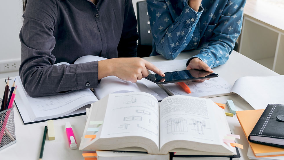 Two people study at a desk with open books and a tablet. One points at the tablet. Papers and pens scattered, a focused mood.
