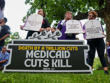 Alt text: A low-angle photograph of protestors on a grassy lawn holding large tombstone-shaped signs. In the foreground, a large black and white sign reads, "DEATH BY A TRILLION CUTS. MEDICAID CUTS KILL." Behind it, three women hold individual tombstone signs reading, "R.I.P. SACRIFICED FOR THE RICH," "SACRIFICED FOR PROFIT," and "R.I.P. HERE LIES AMERICA'S FUTURE."