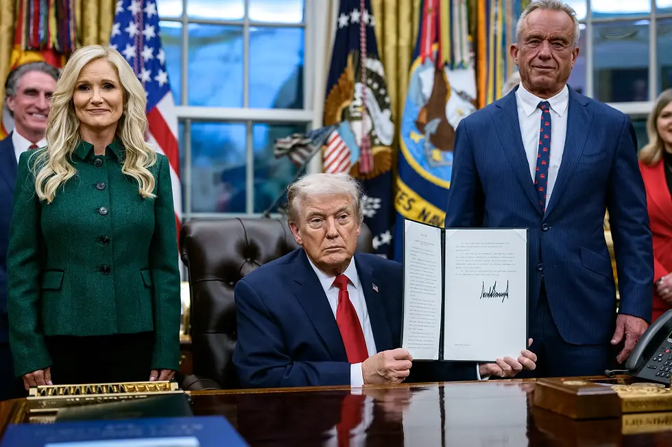 President Trump seated at the Resolute Desk holds up a signed executive order. HHS Secretary Robert F. Kennedy Jr. stands to his right and Kathryn Burgum, Senior Advisor for Addiction Recovery, stands to his left. American flags are visible in the background of the Oval Office.