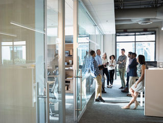 Smiling coworkers chat in a bright modern office hallway with glass walls and cubicles, relaxed and friendly.
