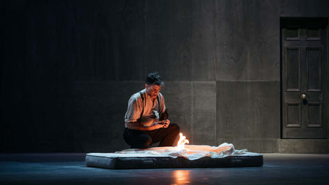 A man lights a small fire on a bare mattress, on a huge, stark stage.