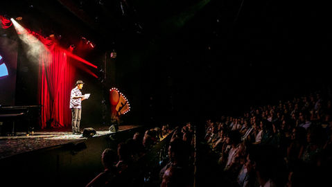 A magician performs at the edge of a red-curtained stage before the large audience.