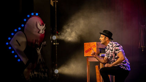 A magician sits beside a box of secrets, as haze billows across the stage.
