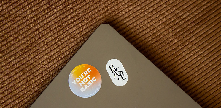 Ordinateur sur fond en velours côtelé avec stickers “YOU'RE NOT BASIC” et “BASE”, symbolisant l’identité visuelle de l’agence