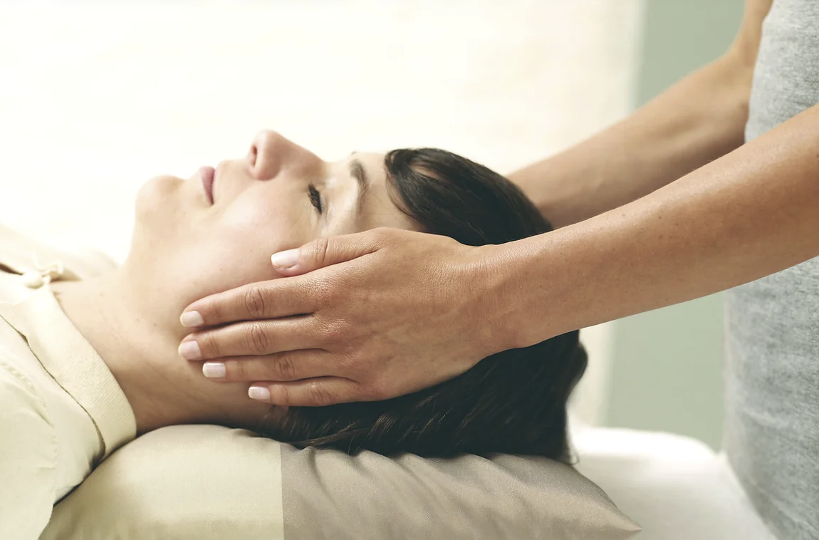 Image of a man lying down, receiving Reiki. Only his head and shoulders are visible. A Reiki practitioner is holding their hands a small distance above her eyes and face, focusing energy on the upper part of her body for healing and relaxation.