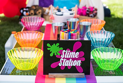Colorful outdoor slime party setup featuring a vibrtant rainbow table runner, neon mixing bowls in pink, orange, yellow, green, and blue, metallic cups, slime-making supplies, and a bold "Slime Station" sign with splatter graphics. Perfect for a sensory activity at a kids birthday party, summer playdate, or creative event for children in Erie, Colorado.