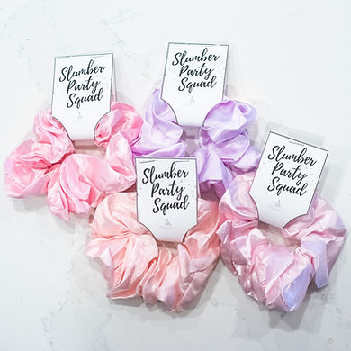Set of pastel pink and purple satin scrunchies with 'Slumber Party Squad' tags — perfect sleepover party favors for girls, pre-teens, and tweens. Customizable add-on for kids spa parties, sleepover setups, and birthday party packages by Katie’s Party Lab in Erie, Colorado.
