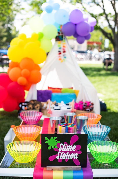 Colorful outdoor slime party setup featuring a rainbow balloon arch over a white teepee tent, vibrant slime-making station with neon mixing bowls, metallic cups, markers, and supplies. A bold "Slime Station" sign sits on a rainbow table runner, with stuffed animals in the background. Perfect for kids birthday parties, pride celebrations, summer festivals, or creative craft party themes.