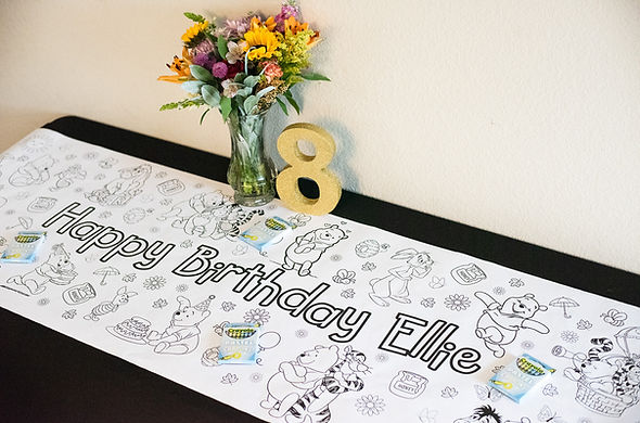 Winnie the Pooh-themed birthday table runner featuring 'Happy Birthday Ellie' message, black-and-white coloring design with classic Pooh characters, floral centerpiece in glass vase, and gold glitter number 8 birthday decor — perfect for a customizable kids birthday party by Katie’s Party Lab in Erie, Colorado.