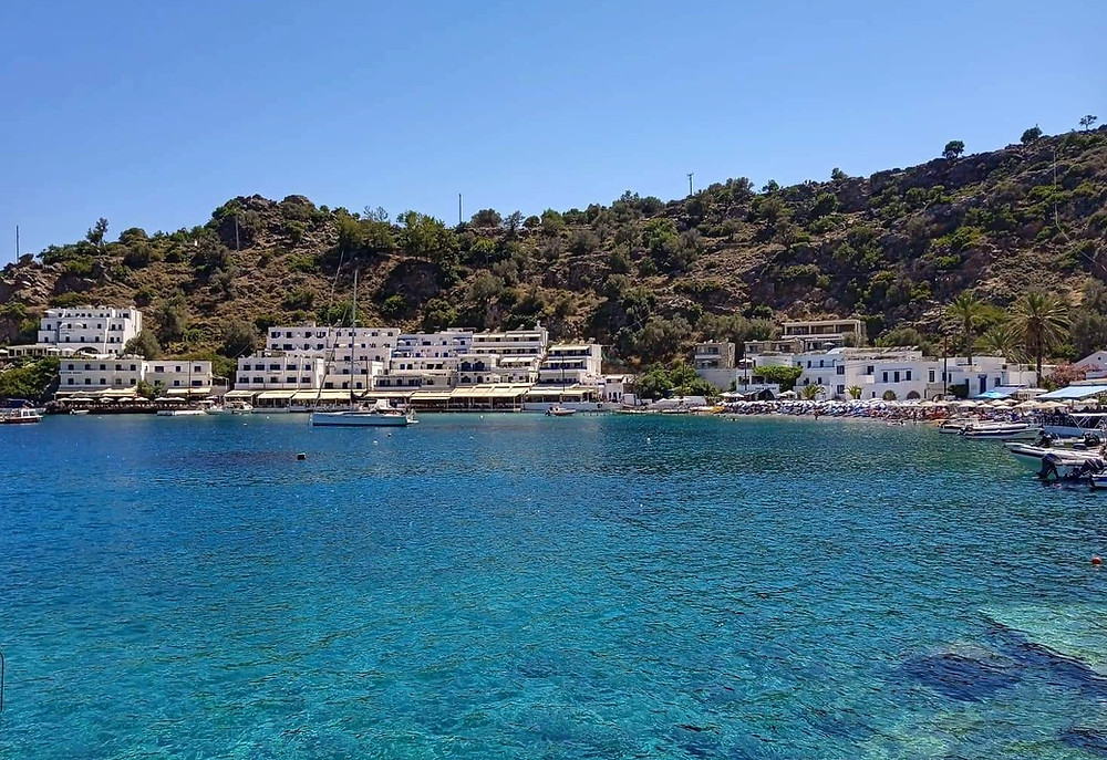 The port of Loutro, with the blue waters, boats, whitewashed buildings, hotels and taverns, as well as the hill above the village, are visible