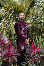 Akash in an embroidered burgundy jacket standing among tropical plants before the Sangeet at his Hindu wedding in Cancun