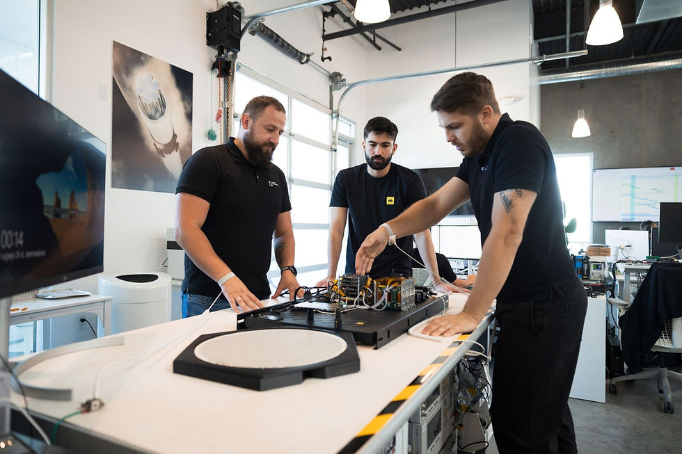 Three men in a tech lab work on electronic equipment. One gestures at a circuit board. Screens and diagrams are visible in the background.