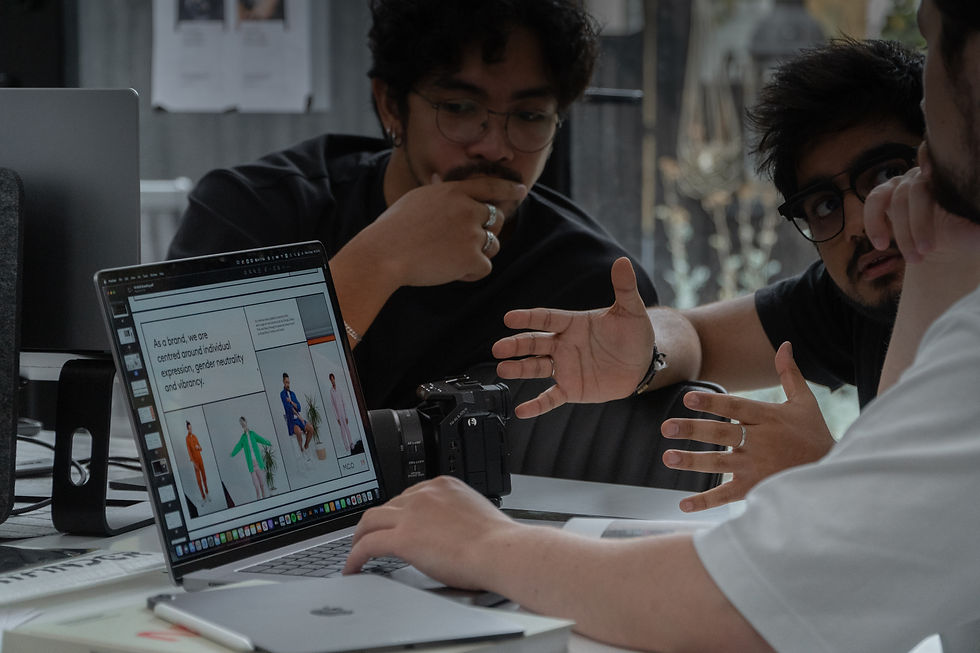 Three people discuss ideas around a laptop displaying fashion designs. Camera and papers on the table, creating a focused, creative mood.