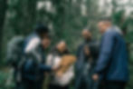 Six people are standing outdoors in a wooded area looking at a map and smiling, dressed for hiking with backpacks.