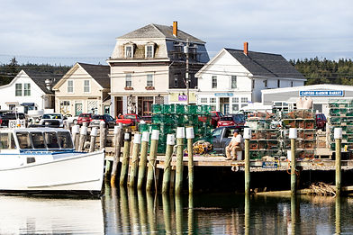 A photo of the docks in Vinalhaven, Maine. The photo includes a docked boat, a pier with lobster cages and a person sitting on it, and commercial buildings in the background