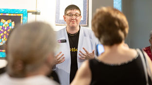 Person with glasses and name tag Sarah Busman speaks to an audience in an art gallery with colorful quilts, hands raised.