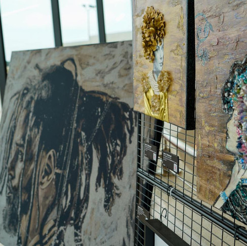 A display of several artworks. On the right, two mixed-media paintings feature profile portraits adorned with three-dimensional embellishments like gold leaf, beads, and textured materials. To the left, a large, dimly lit canvas shows a portrait of a person with dreadlocks (J Cole) in a grayscale palette.