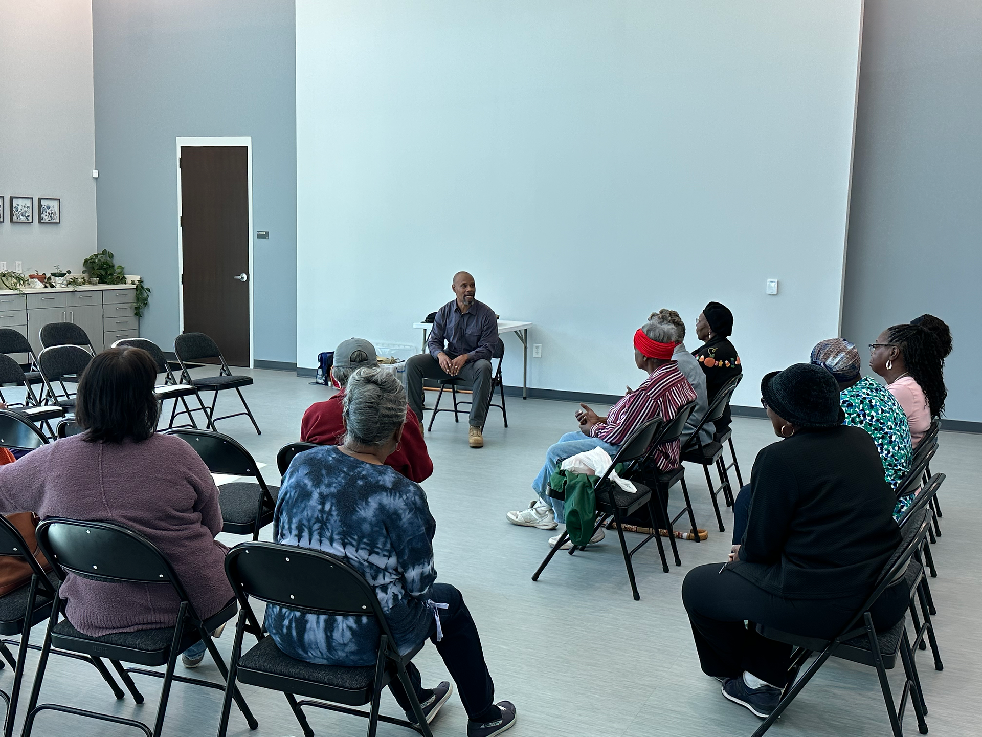 An indoor photo of a group of people sitting in black folding chairs, arranged in a semi-circle, listening to a man seated on a chair in the center of the room. The room has light gray walls, a light-colored floor, and a large white door to the left. A small table with a water bottle and cup is next to the speaker. The attendees are casually dressed, facing the speaker, and appear to be engaged in a presentation or discussion.