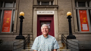 Smiling man (Robert Pinson) stands in front of brick building labeled "The Arts Center," with red banners, round lights, and steps leading to entrance.