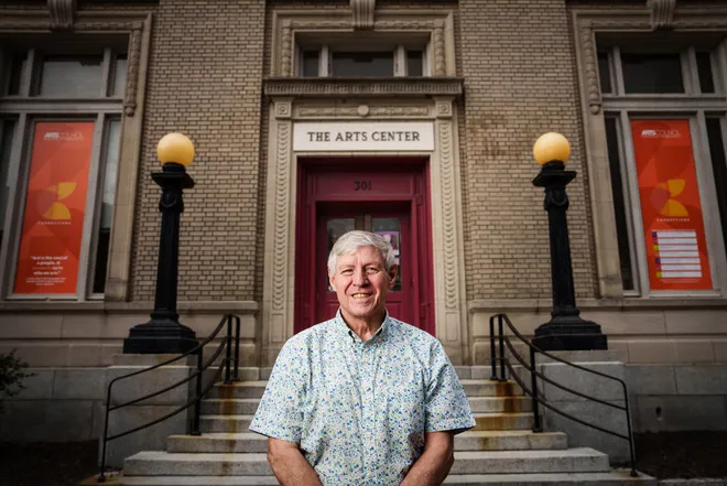 Smiling man (Robert Pinson) stands in front of brick building labeled "The Arts Center," with red banners, round lights, and steps leading to entrance.