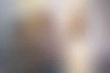 A man with a reddish-brown beard and a woman with her hair pulled back are standing side-by-side, looking at photographs displayed on a light-colored wall at an art exhibition. The man is wearing a tan quarter-zip fleece, and the woman is wearing a striped green and beige sweater.