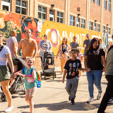 A diverse crowd of people walking on a sunny day in front of a red brick building with a large orange banner that says "COMMUNITY." The banner features abstract faces and colors.