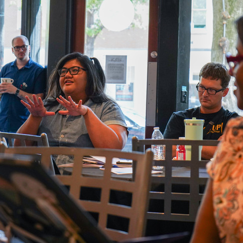 A medium shot of a group of people sitting around tables in a brightly lit indoor space, likely a café or office meeting area. In the center, a woman with glasses and long dark hair gestures with both hands open while speaking. To her right, a man in a black t-shirt and glasses listens intently. In the background, a man stands near a large window holding a white mug. In the foreground, another woman is partially visible from the side, and a laptop screen is blurred in the lower left corner.