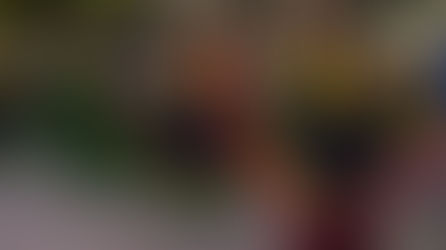 A group of four women are dancing in a street parade. They are wearing black dresses with ruffles of red, yellow, and blue, the colors of the Colombian flag, on their skirts. The ruffles on their sleeves are green, yellow, and red. They also have colorful flower headbands and are smiling as they perform. Their arms are extended out to the sides, holding the edges of their full skirts, which they have spread out wide as they move. The street is lined with trees and buildings, and there are people watching the parade in the background.