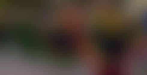 Three women in vibrant, colorful folk dresses are dancing on a city street. Their dresses are black with red, yellow, and blue ruffles. They are holding out the skirts of their dresses as they dance, with a crowd of people watching in the background.